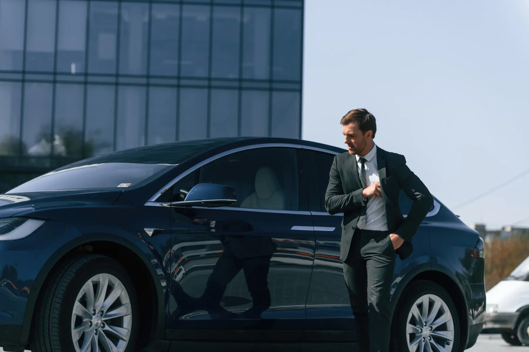 Standing, in black suit. Businessman is near his car outdoors
