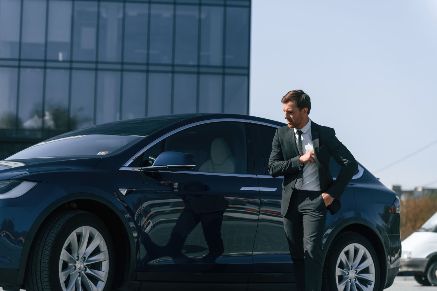 Standing, in black suit. Businessman is near his car outdoors